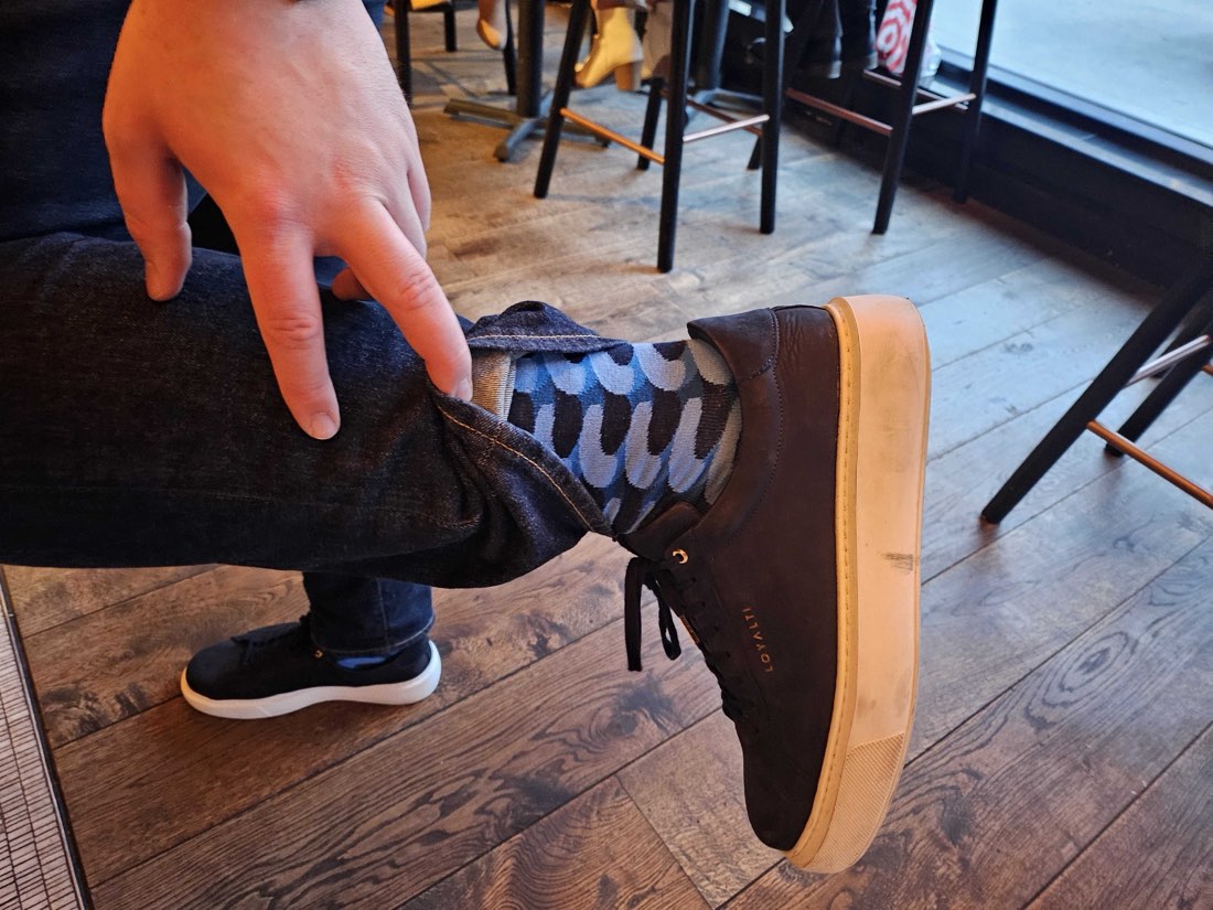 Someone on a bar stool pulling a jean cuff up over a brown leather high-top to show a blue patterned Rovertown sock, on a wood floor