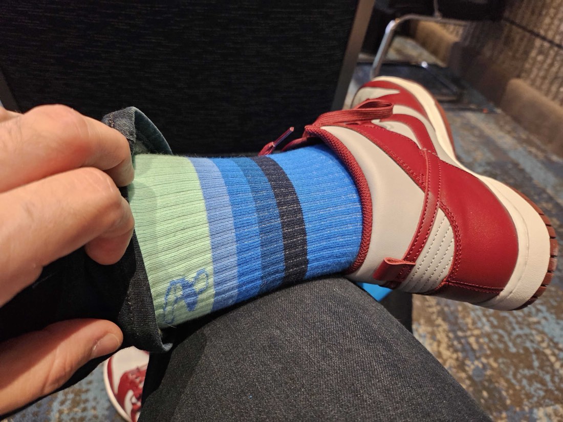 A hand pulling a trouser cuff back to reveal a mint and blue striped Rovertown sock above a red and white sneaker in a conference chair