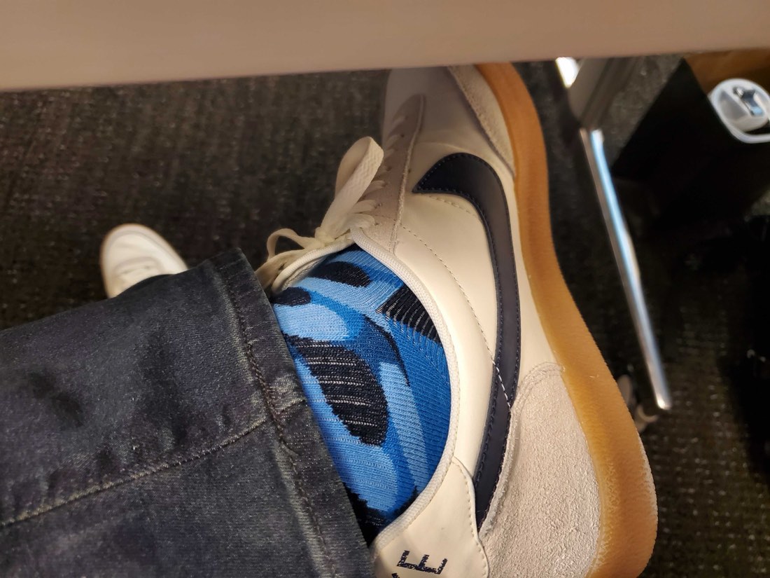 An ankle crossed under a desk in a white leather Nike sneaker with a gum sole, showing a bright blue patterned Rovertown sock