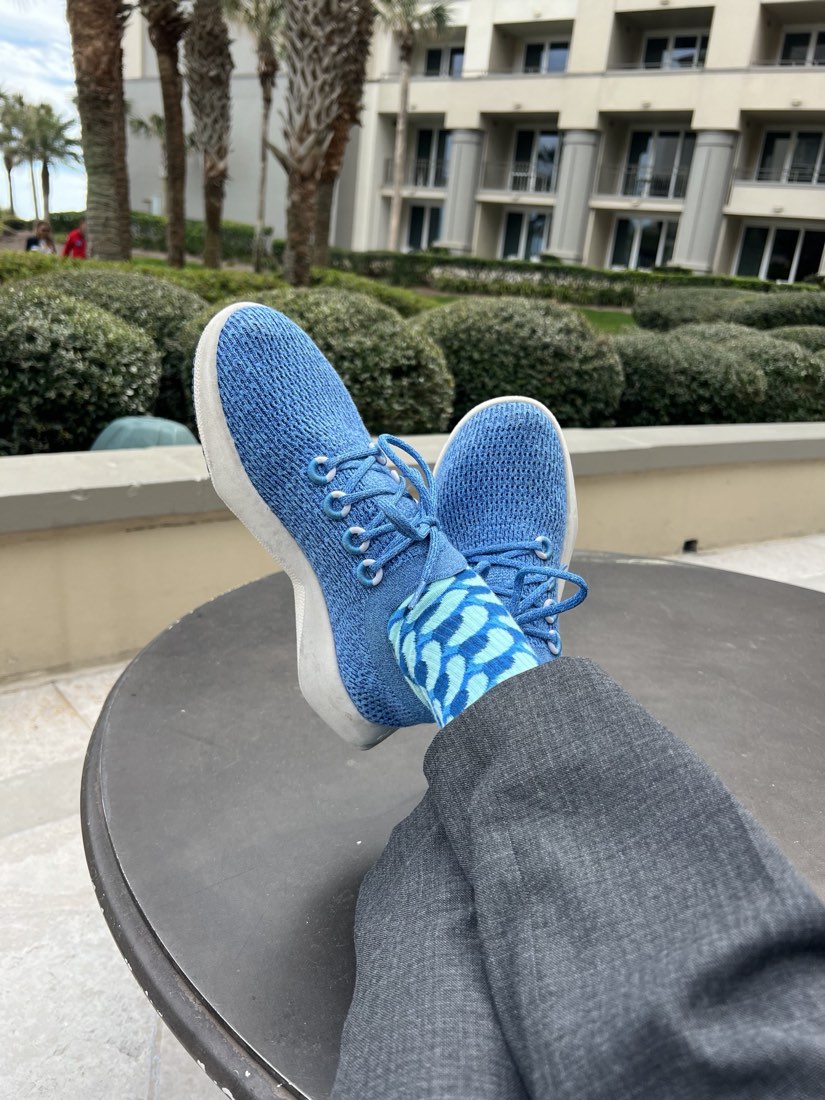Legs stretched across a patio table in front of palm trees and a resort building, wearing blue knit sneakers and tie-dye blue Rovertown socks