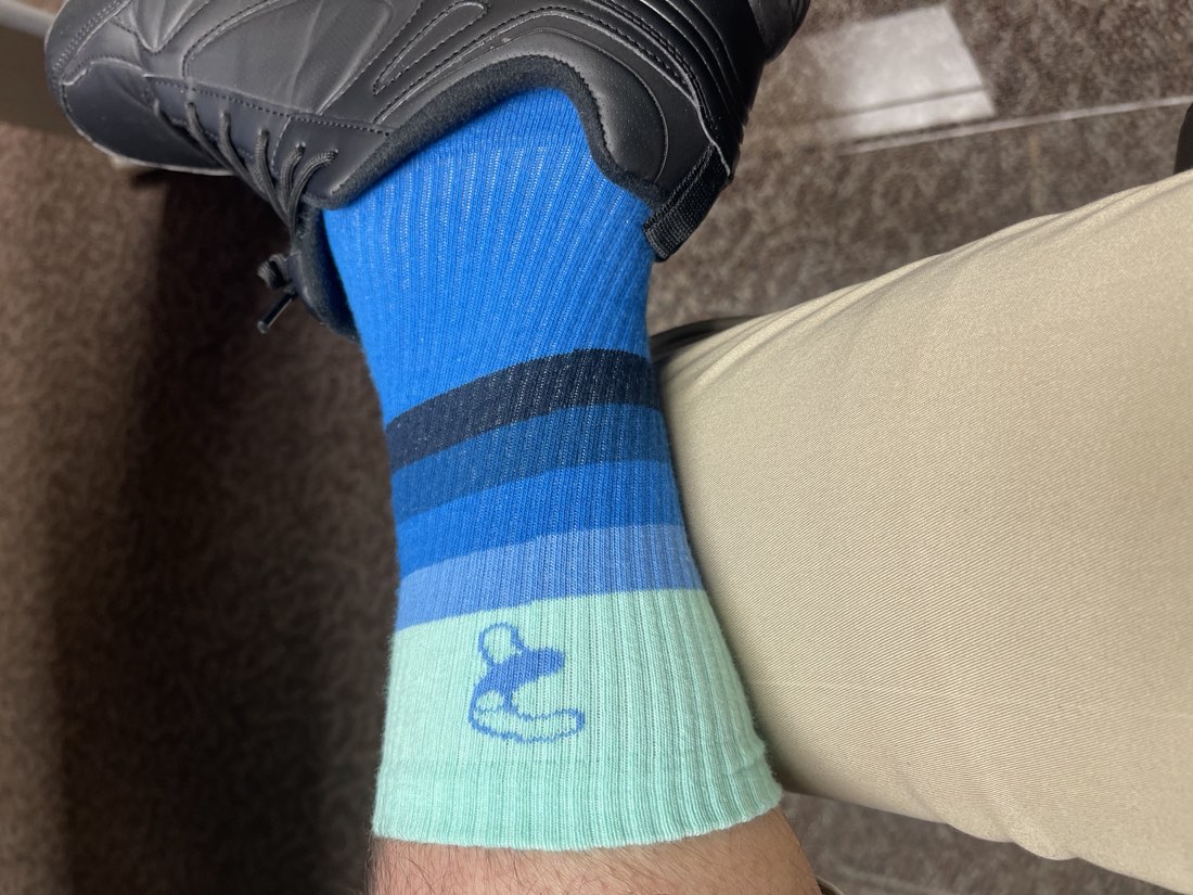 Close-up of an ankle in a mint and blue striped Rovertown sock carrying the Rovertown mark, above a black dress shoe