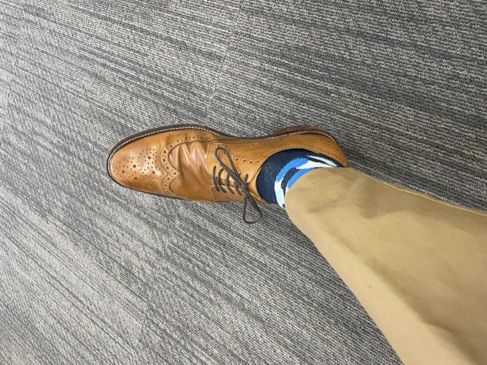 Tan leather wingtip brogue and khaki trousers on gray office carpet, with a blue Rovertown sock at the ankle