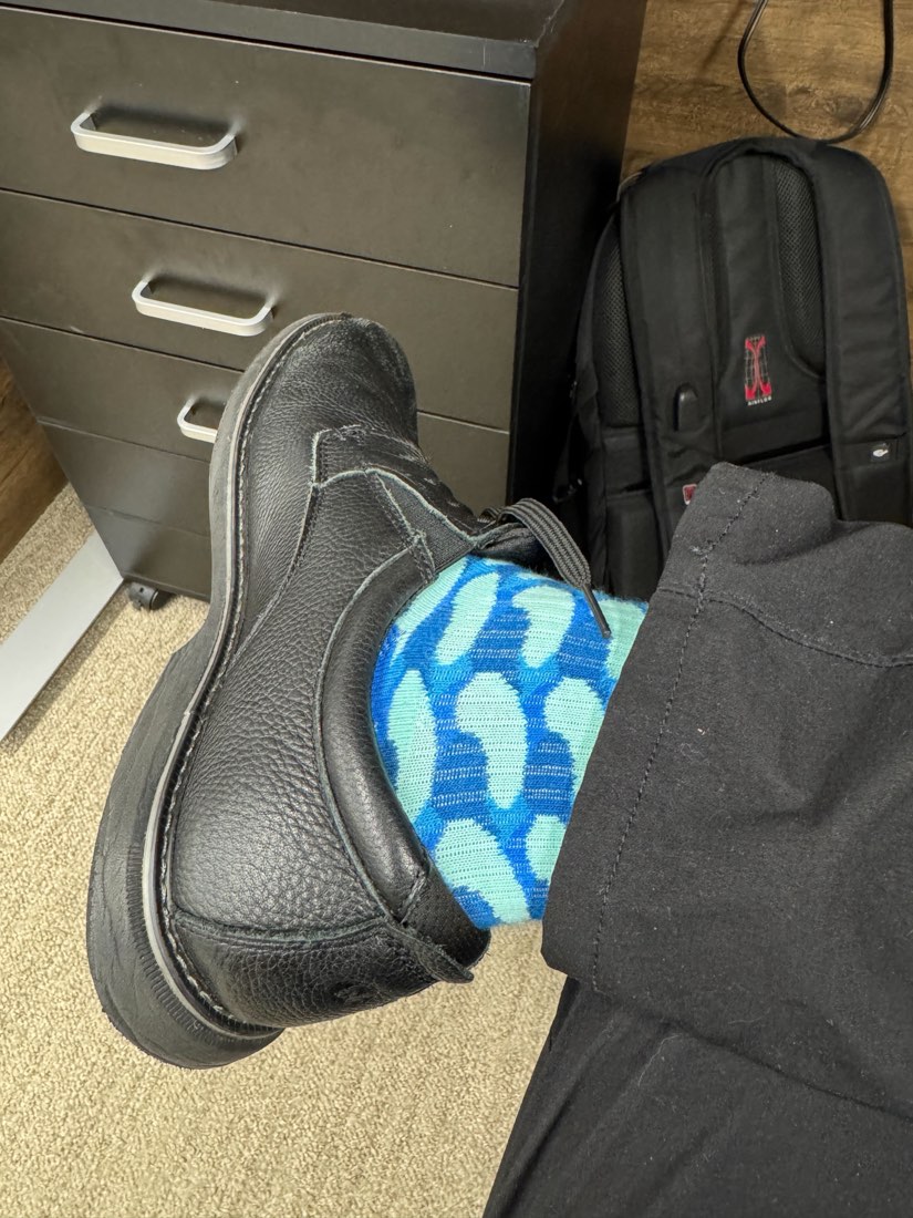 A black leather work boot propped beside a filing cabinet and a backpack, showing an aqua and blue patterned Rovertown sock